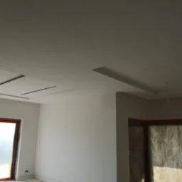 Wnętrze w trakcie wykańczania, z widocznymi elementami sufitu podwieszanego z płyt kartonowo-gipsowych oraz przygotowanymi do malowania ścianami i oknami zabezpieczonymi folią.