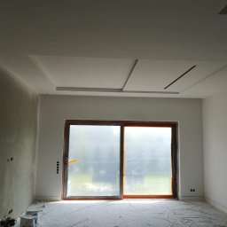 PHU COMPLEX GRZEGORZ ZIMNIAK - Wnętrze pokoju w trakcie remontu, ściany pokryte szpachlą, okno zabezpieczone folią, widoczne puszki z farbą i narzędzia malarskie.