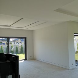 PHU COMPLEX GRZEGORZ ZIMNIAK - Wnętrze w stanie deweloperskim z widocznymi gładkimi ścianami, betonową posadzką i oknami na ogród, z fragmentem kominka po lewej stronie.