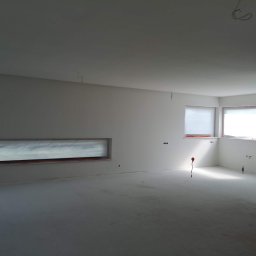 PHU COMPLEX GRZEGORZ ZIMNIAK - Wnętrze w trakcie prac wykończeniowych: gładkie, białe ściany, przygotowane do malowania, widoczne punkty elektryczne i instalacje hydrauliczne, zakryte okno poziome, surowa podłoga.