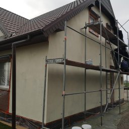 PHU COMPLEX GRZEGORZ ZIMNIAK - Ocieplanie domu jednorodzinnego: ekipa na rusztowaniu aplikuje warstwę izolacyjną na elewację, okna zabezpieczone folią, widoczne wiaderka z materiałem i brązowa dachówka.