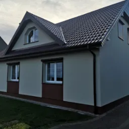 Wykończony dom jednorodzinny z brązowym dachem i jasną elewacją, widok z boku na tle pochmurnego nieba.