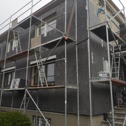 PHU COMPLEX GRZEGORZ ZIMNIAK - Budynek dwukondygnacyjny w trakcie ocieplania, pokryty grafitowym styropianem, otoczony rusztowaniem. Na rusztowaniu widoczne paczki styropianu Fasada Grafit oraz wiadro z zaprawą. Część elewacji...