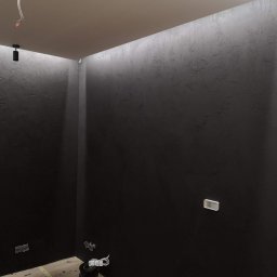 PHU COMPLEX GRZEGORZ ZIMNIAK - Ciemnoszara ściana z dekoracyjną fakturą i ukrytym oświetleniem LED wzdłuż sufitu, widoczne gniazdka elektryczne i puszka z narzędziami na podłodze.