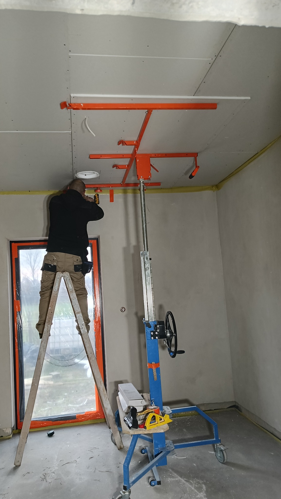 Montaż płyty gipsowej na suficie za pomocą podnośnika. Pracownik na drabinie przykręca płytę. Widoczne narzędzia i fragment okna. Żółta taśma na łączeniu ściany z sufitem.