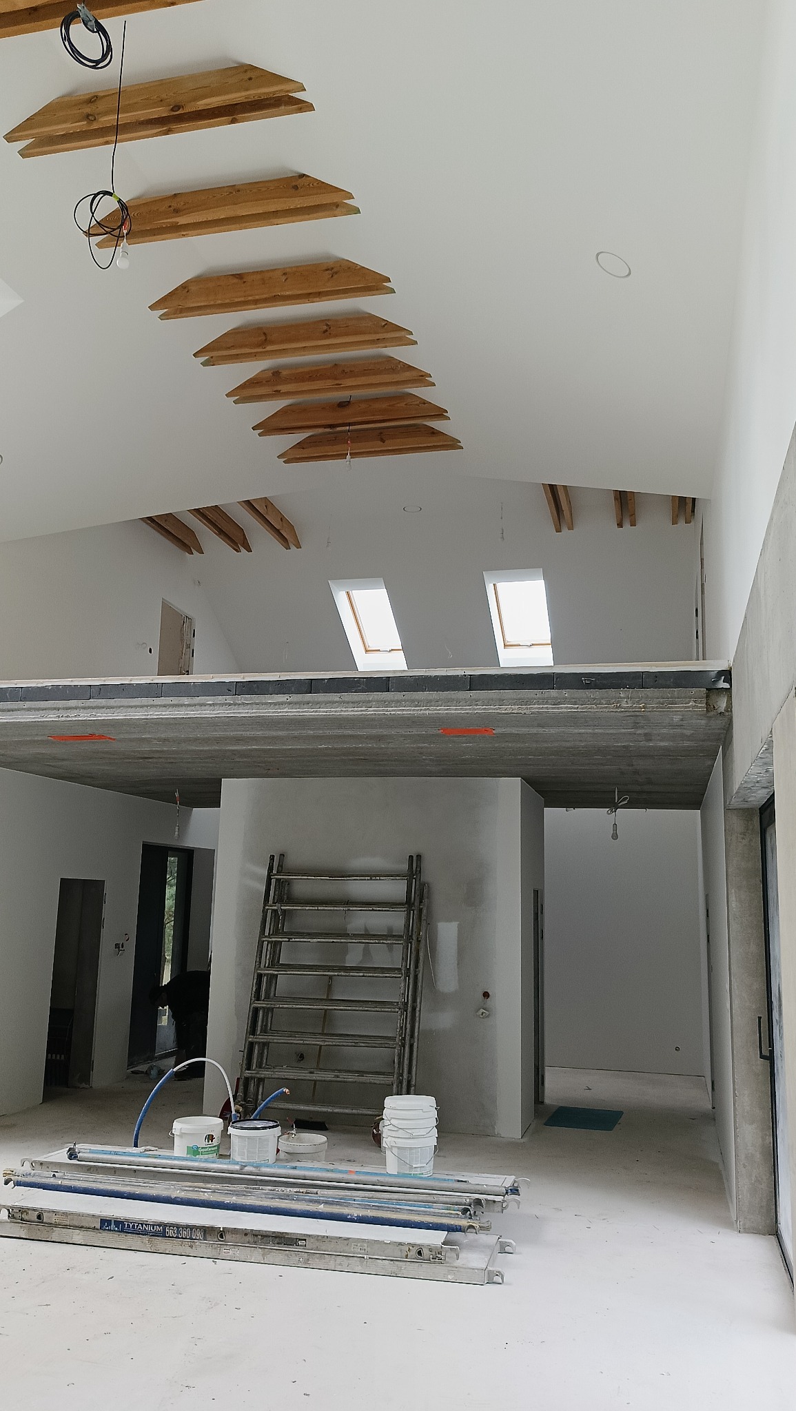 Wnętrze w trakcie prac wykończeniowych: surowy strop, drewniane elementy dekoracyjne pod sufitem, drabina i akcesoria malarskie na podłodze.