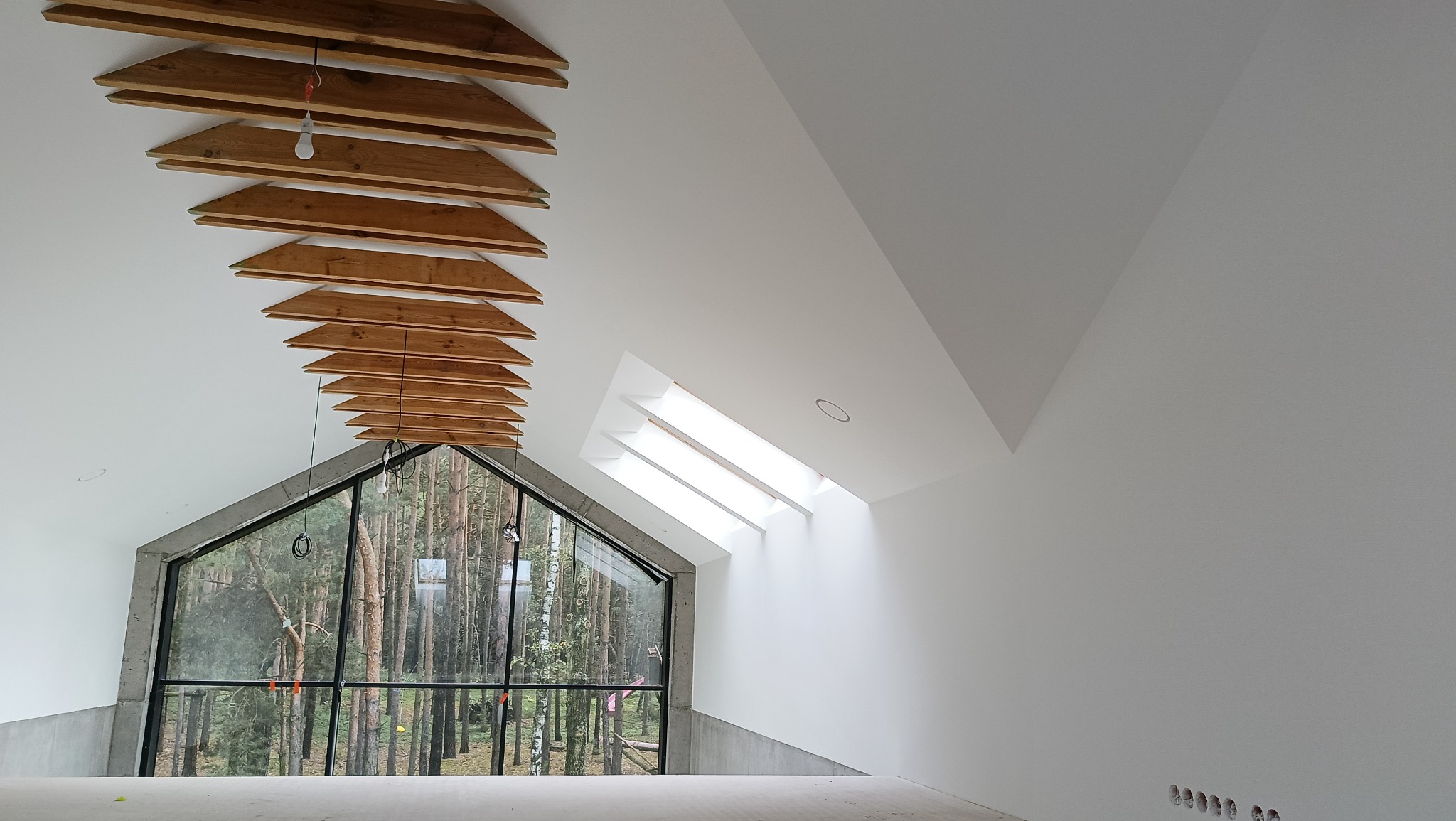 Wnętrze z dużym oknem i drewnianą konstrukcją sufitu. Widoczne gładkie, białe ściany i okno z widokiem na las. Surowy stan wykończenia.