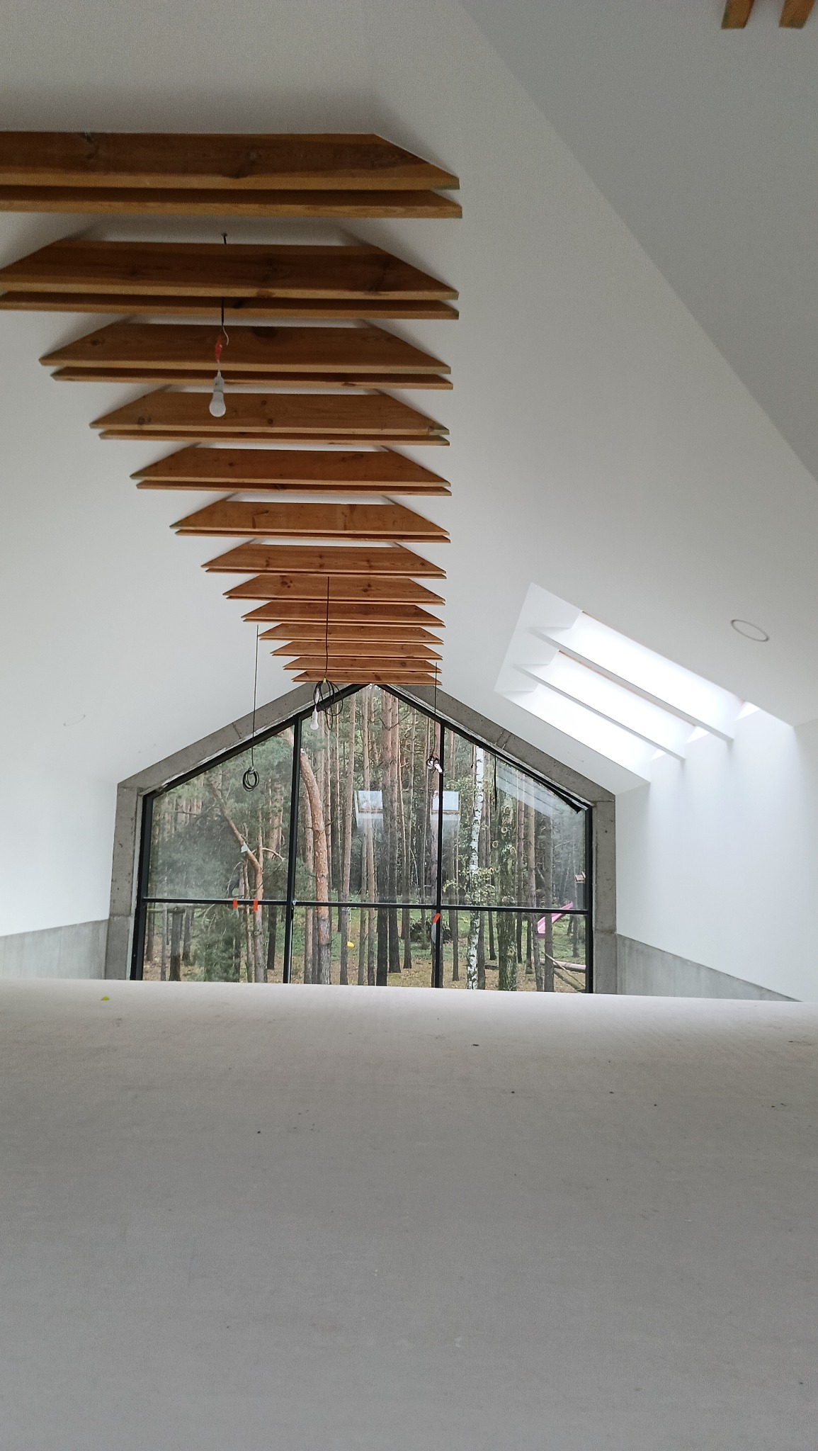 Wnętrze poddasza z oknem szczytowym i drewnianą dekoracją sufitu. Widok na las przez okno. Ściany pomalowane na biało, sufit biały z drewnianymi elementami.