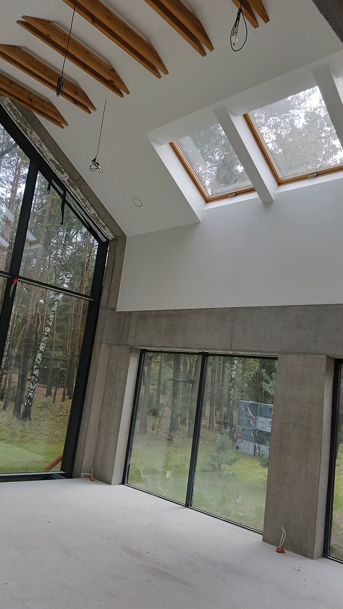 Surowe wnętrze domu z dużymi oknami i drewnianą konstrukcją dachu. Widoczne betonowe elementy i wpadające światło dzienne. Minimalistyczny design w trakcie wykończenia.
