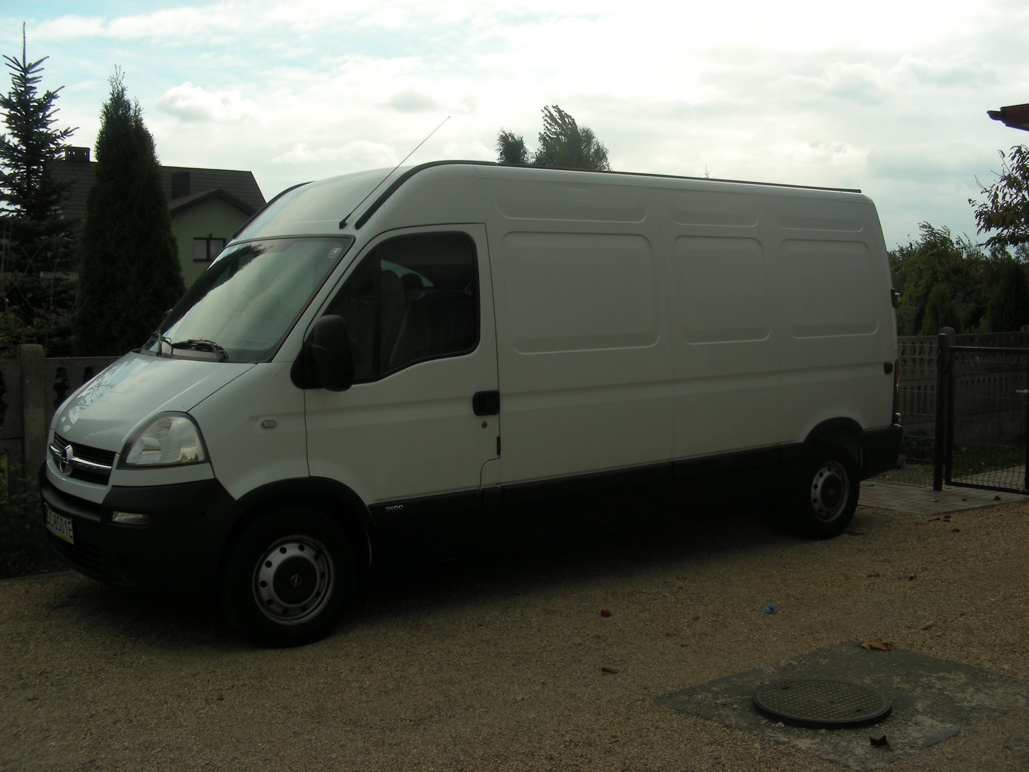 Biały bus dostawczy Opel Movano zaparkowany na żwirowym podjeździe, z widoczną anteną na dachu i czarnym pasem ochronnym wzdłuż dolnej części nadwozia.