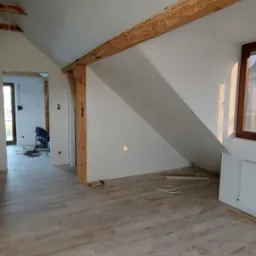 Poddasze w trakcie wykańczania: położona podłoga z paneli imitujących drewno, ściany pomalowane na biało, widoczne drewniane belki stropowe i fragment grzejnika.