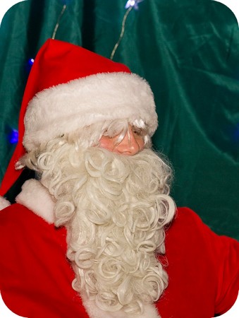 Portret osoby przebranej za Świętego Mikołaja, z białą brodą i czerwonym strojem na tle zielonej zasłony z delikatnymi światełkami.