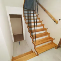 Jasne drewniane schody z czarną, metalową balustradą w trakcie montażu, widoczne otwory na ścianie i zabezpieczona poręcz, prowadzące do niezakończonego pomieszczenia.