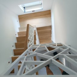 Stolarstwo Marszałek Przemysław - Nowoczesne schody drewniane z białą, geometryczną balustradą, widziane z góry. Naturalne światło z okna dachowego oświetla drewniane stopnie.