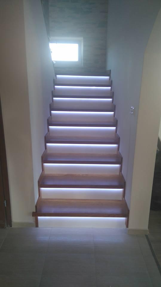 Drewniane schody z podświetleniem LED na każdym stopniu, widok od dołu, jasne światło z okna na górze, szare płytki podłogowe.