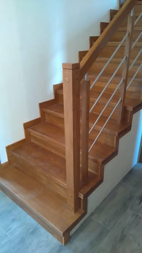 Drewniane schody z balustradą ze stali nierdzewnej, widoczne z dołu, z drewnianymi słupkami i poręczą, na tle białej ściany i szarej podłogi.