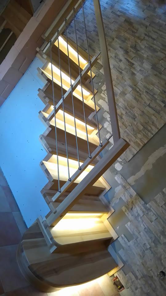 Drewniane schody z podświetleniem stopni i balustradą z cienkich, pionowych, nierdzewnych prętów, widok od dołu, ściana wykończona kamieniem dekoracyjnym.