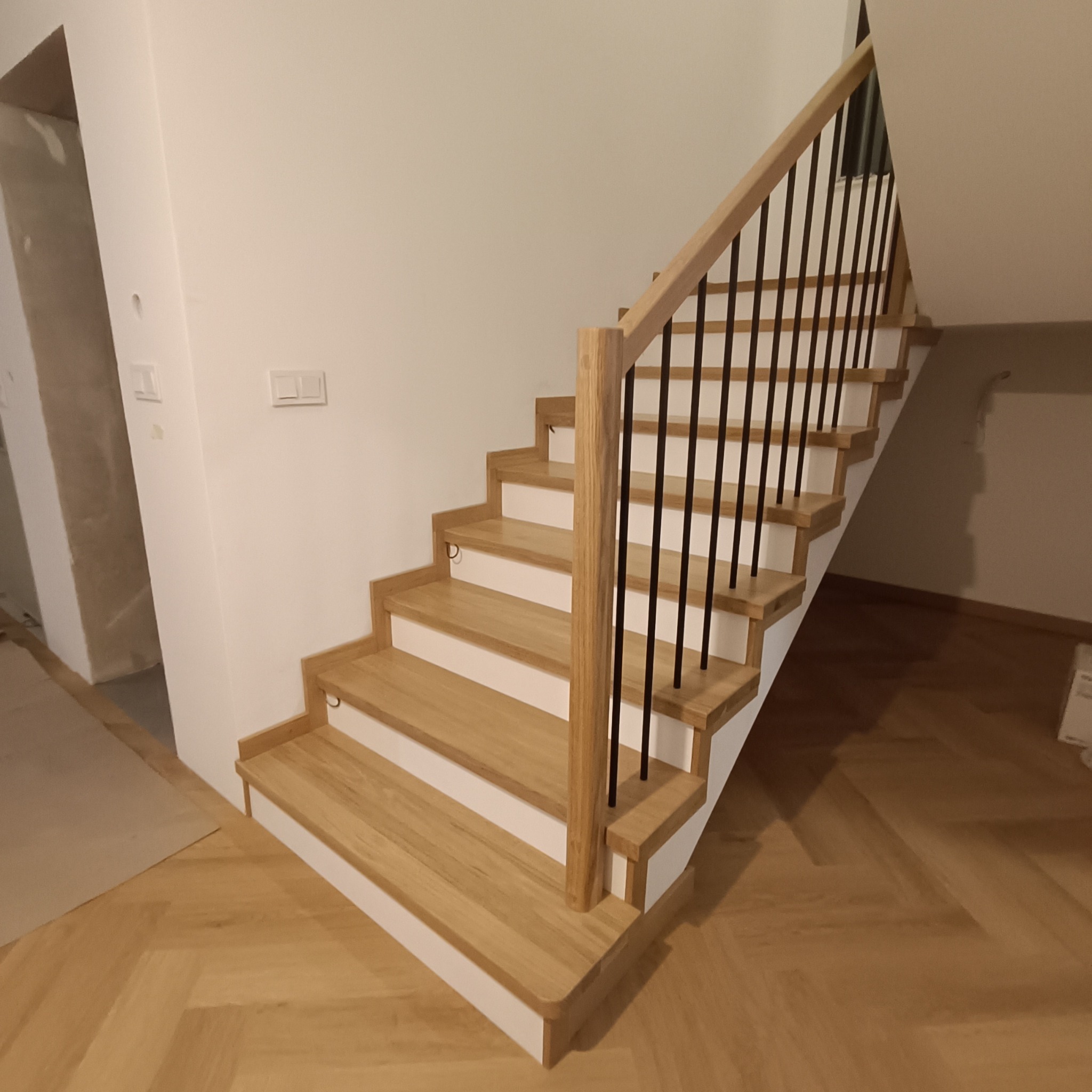Jasne, dębowe schody z czarnymi, metalowymi prętami balustrady i białymi podstopnicami, widziane z dołu, na tle białej ściany i parkietu w jodełkę.