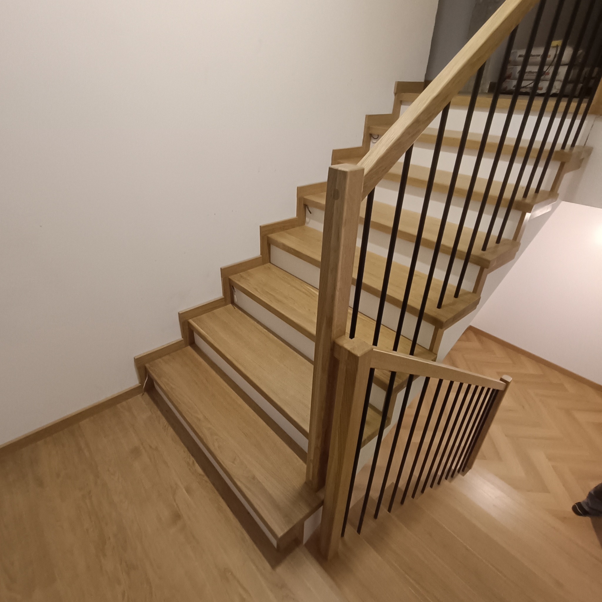 Jasne, drewniane schody z czarnymi, metalowymi prętami balustrady, widziane z góry. Parkiet w jodełkę na podłodze. Minimalistyczny design.