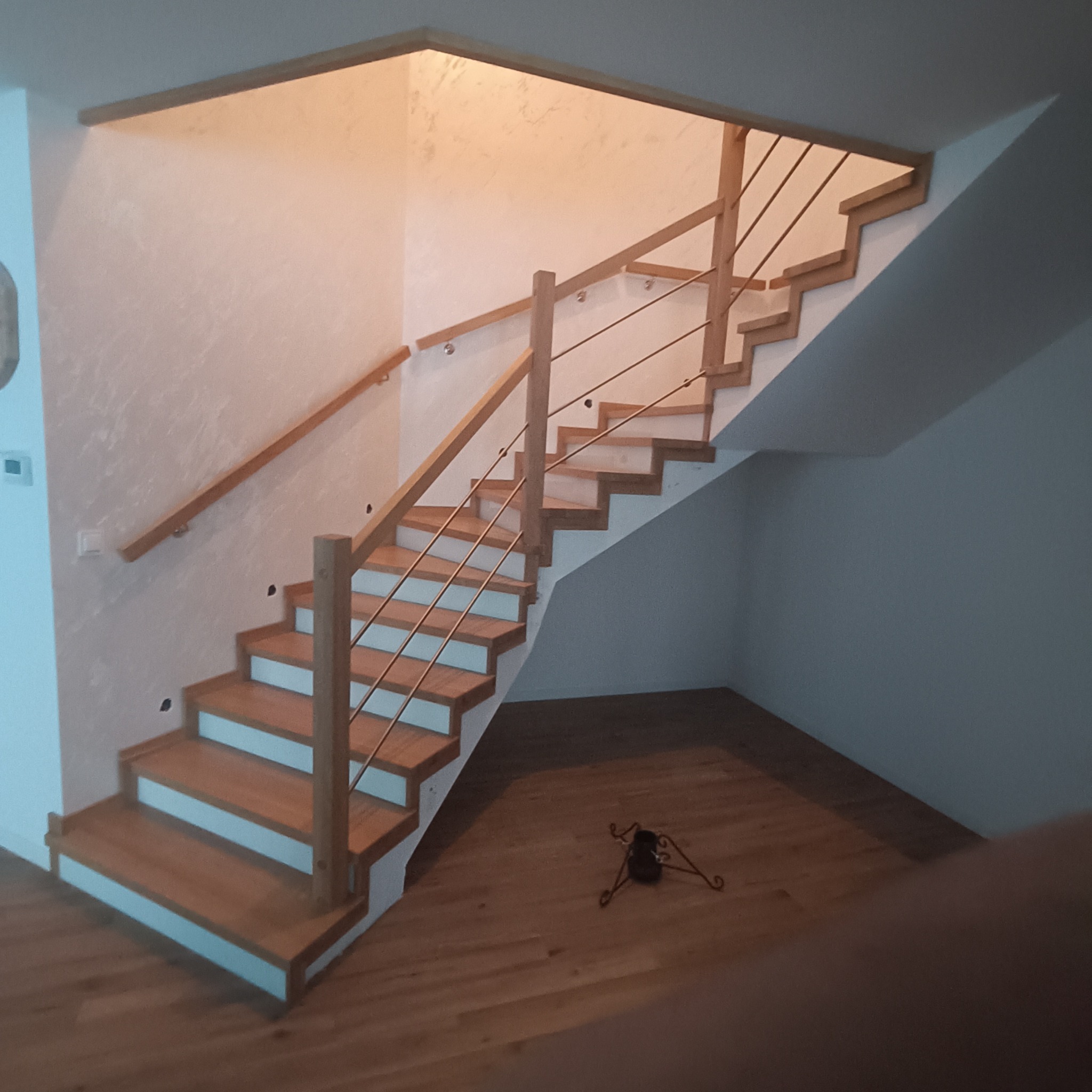 Jasne, drewniane schody z metalową balustradą w nowoczesnym wnętrzu. Widoczne oświetlenie górne i drewniana podłoga. Minimalistyczny design.