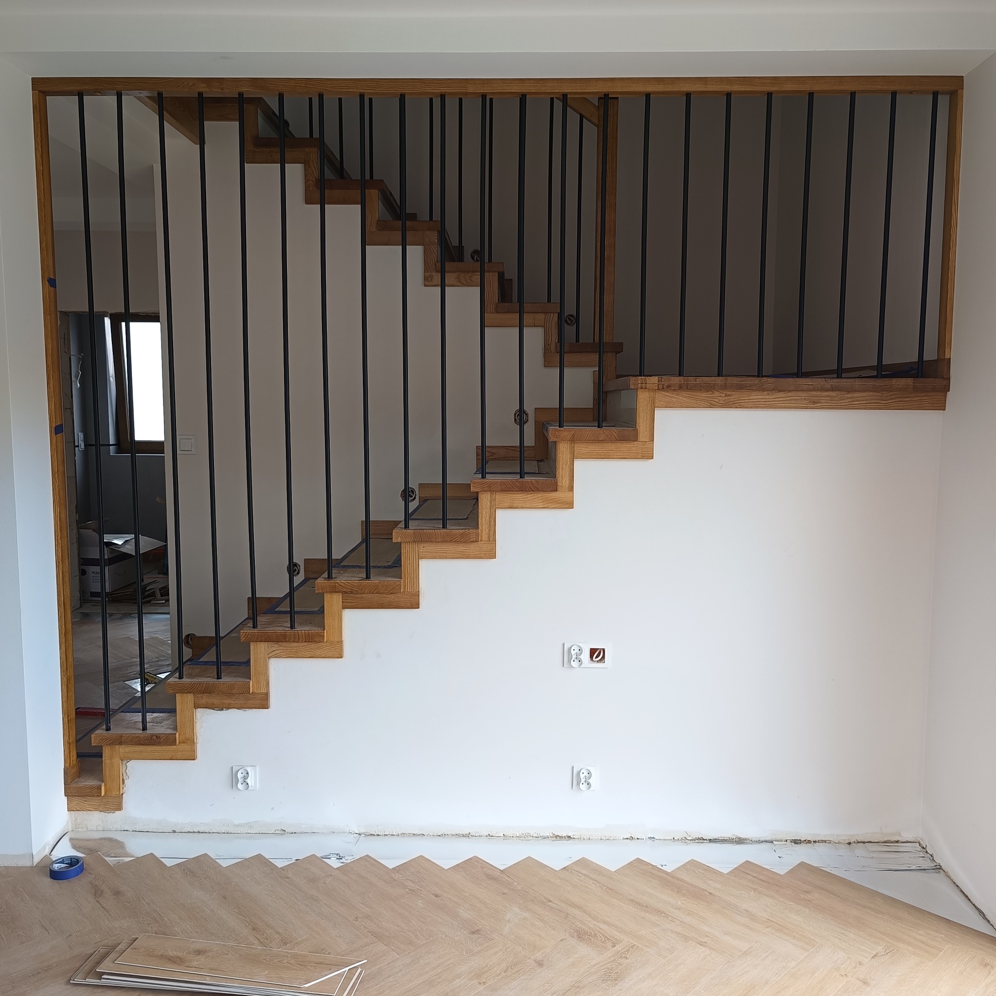Montaż dębowych schodów z czarną balustradą sztabową. Widoczne przygotowane podłoże pod parkiet i gniazdka elektryczne na ścianie.
