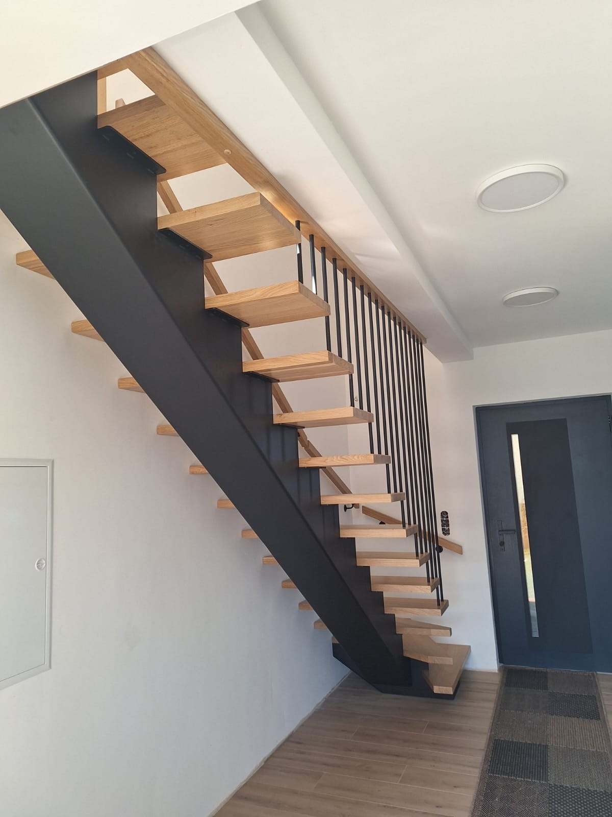 Nowoczesne schody z drewnianymi stopniami i czarną, metalową konstrukcją nośną oraz balustradą z pionowych prętów. Widok z dołu, z boku, na tle białej ściany i drzwi.