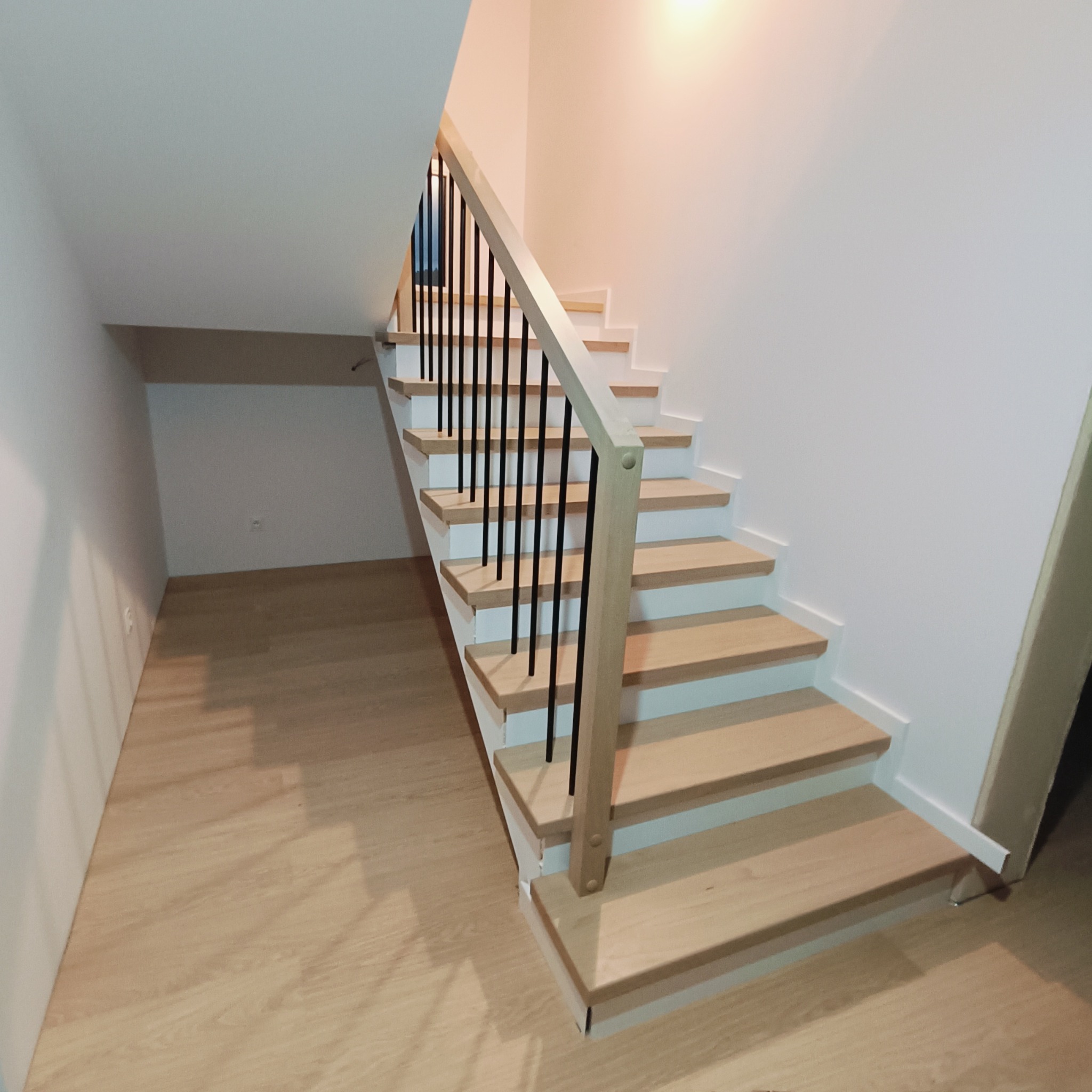 Jasne, drewniane schody z czarnymi, metalowymi prętami balustrady w nowoczesnym wnętrzu. Minimalistyczny design, naturalne światło, widok z dołu na górę.