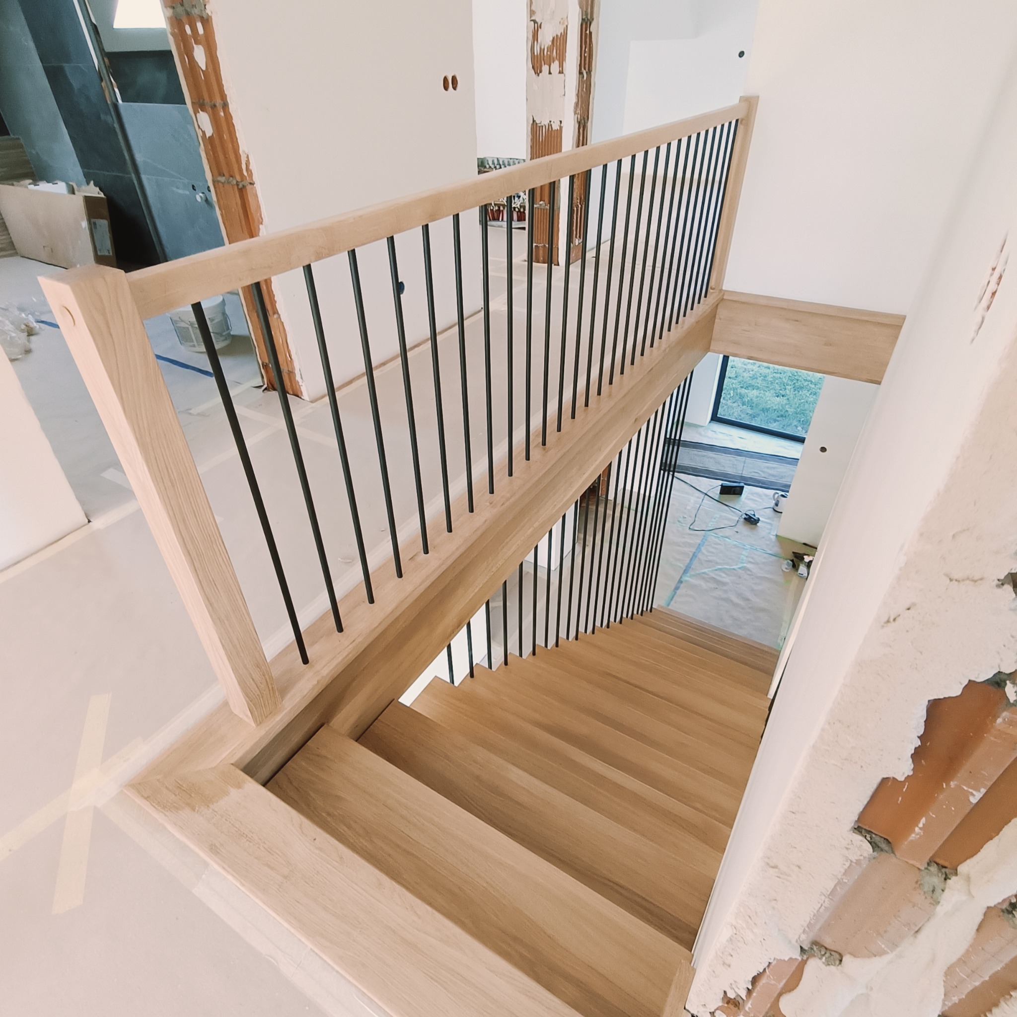 Jasne, drewniane schody z czarną, metalową balustradą w nowoczesnym wnętrzu w trakcie wykończenia. Widok z góry na dół, z fragmentami surowych ścian.