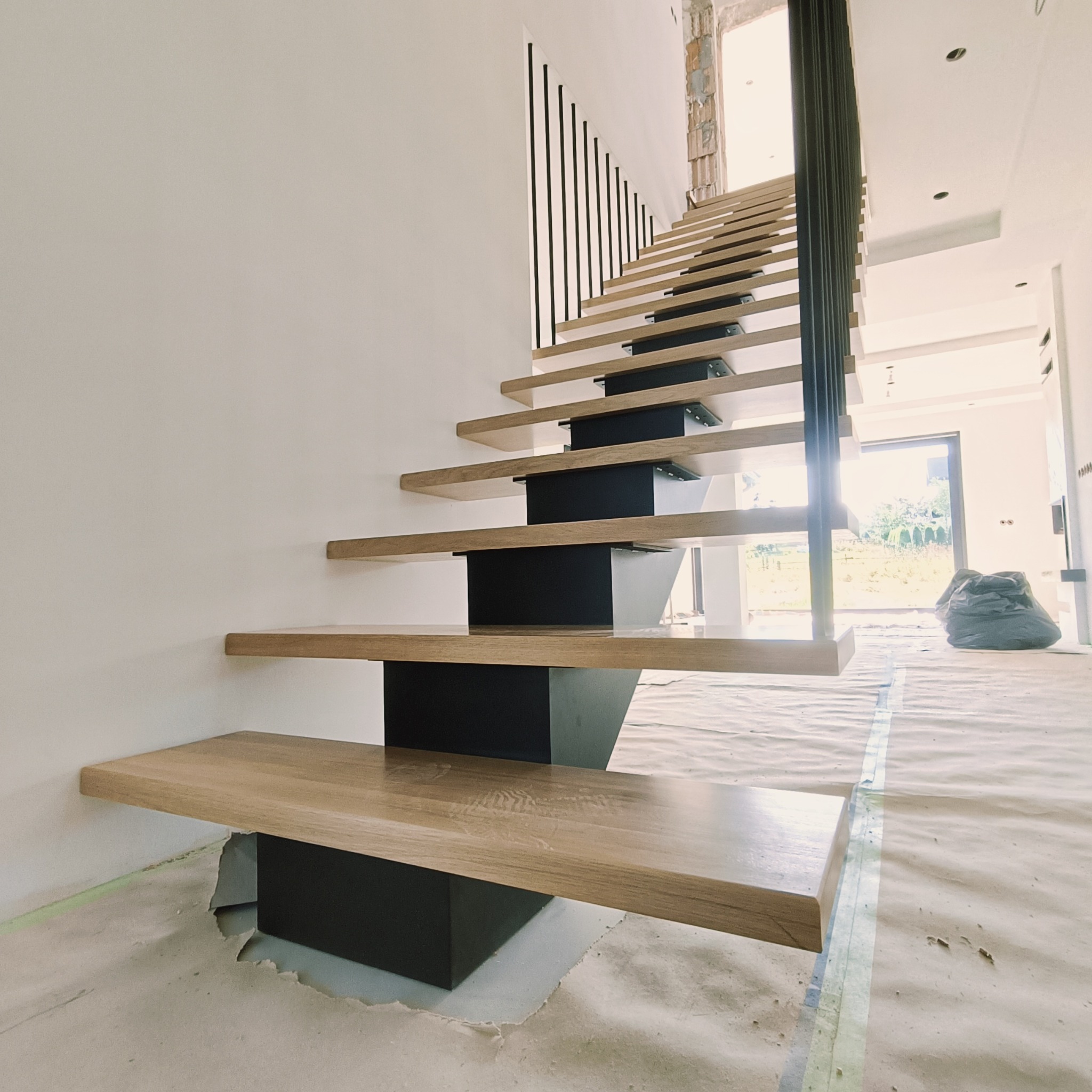 Nowoczesne schody z drewnianymi stopniami i czarną balustradą, widok z dołu, w tle okno i elementy wykończeniowe wnętrza.