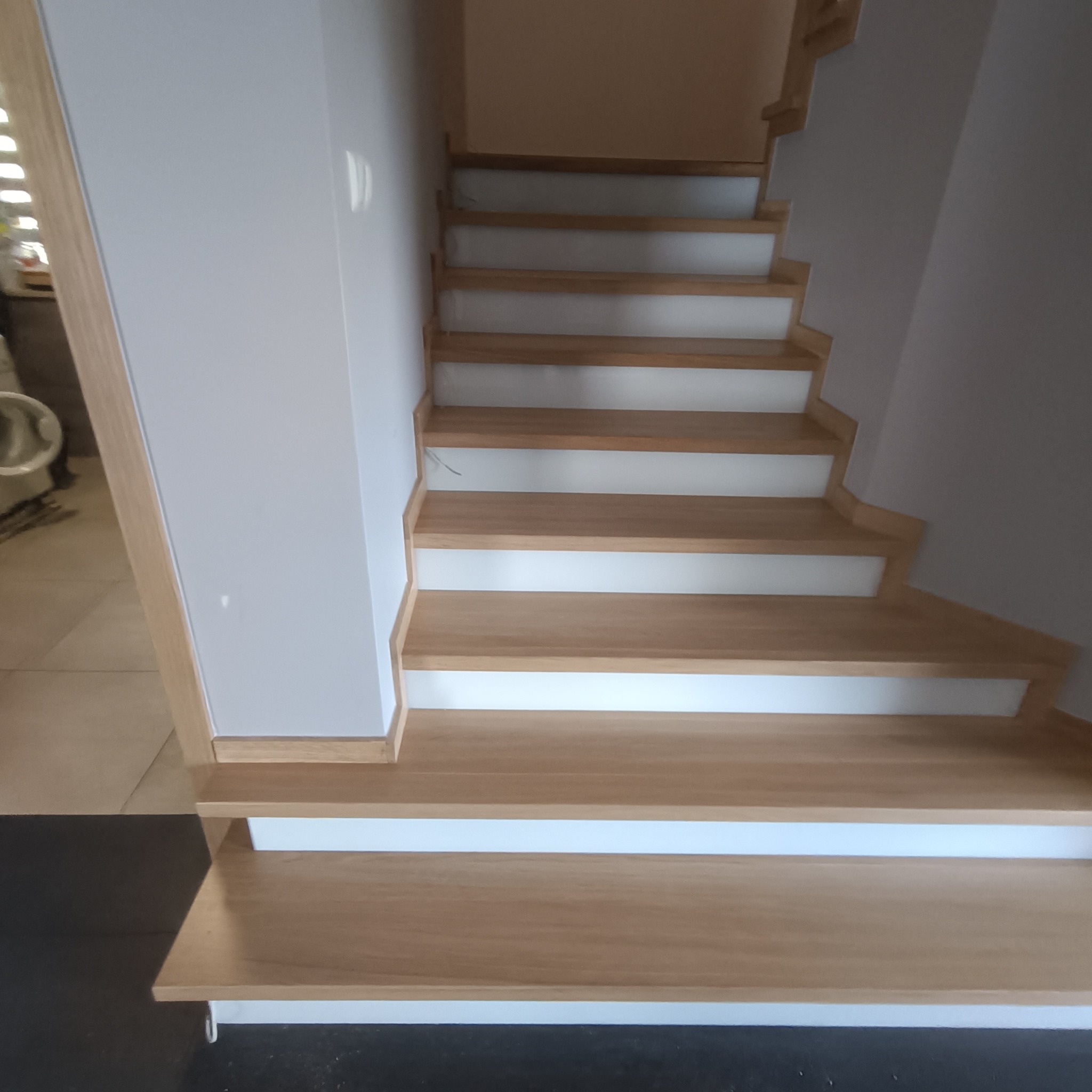 Jasne, drewniane schody z białymi podstopnicami, widziane z dołu. Minimalistyczny design, naturalne oświetlenie, widoczne detale wykończenia.