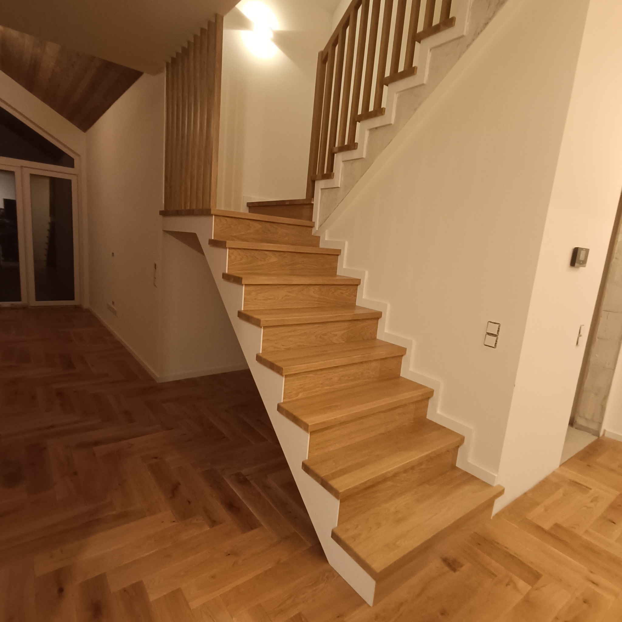 Jasne, dębowe schody z białymi podstopnicami i drewnianą balustradą w nowoczesnym wnętrzu. Parkiet ułożony w jodełkę. Minimalistyczny design z akcentami naturalnego drewna.