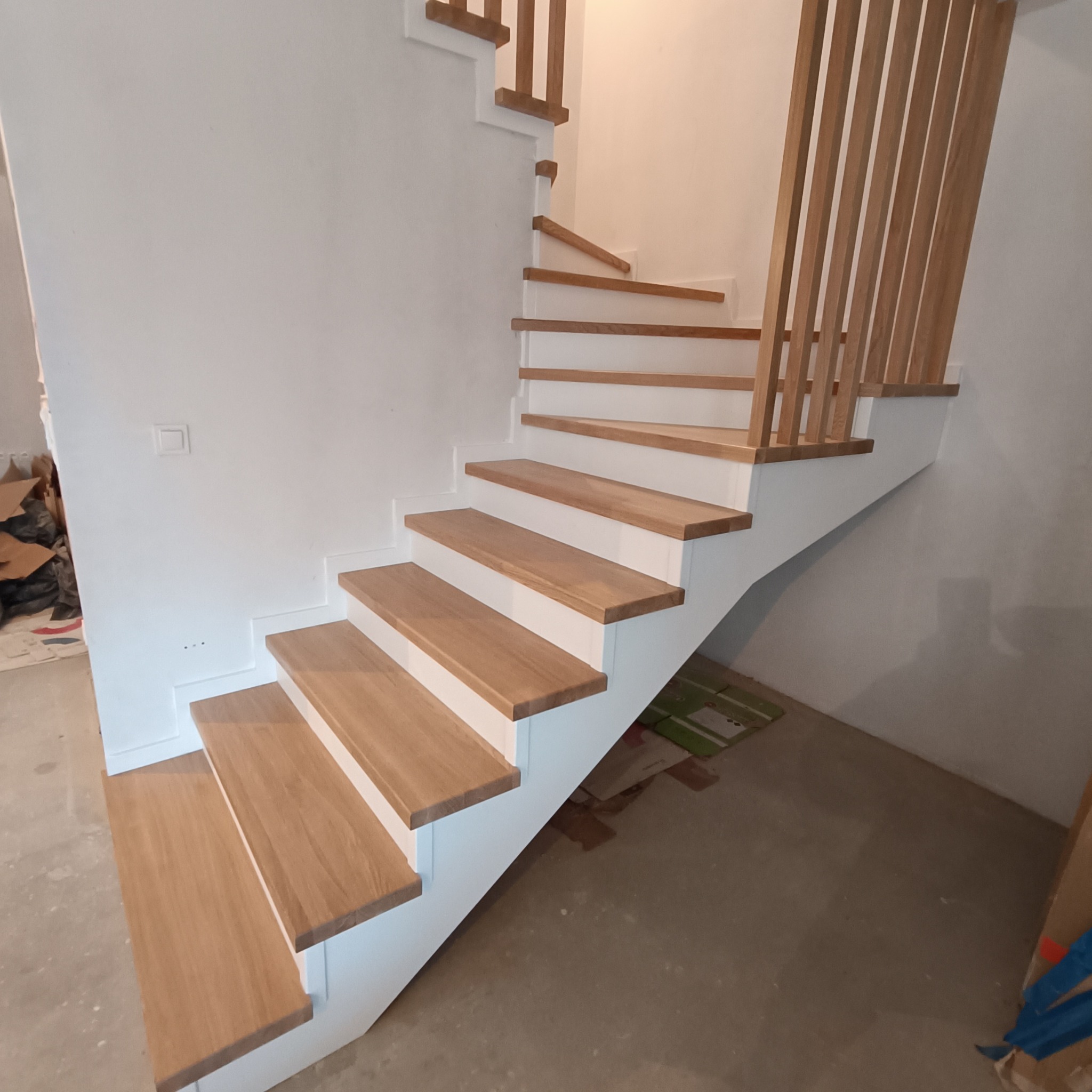 Drewniane schody z białymi podstopnicami i ażurową balustradą z pionowych listew. Widok z dołu, ukazujący zakręt schodów i minimalistyczny design.