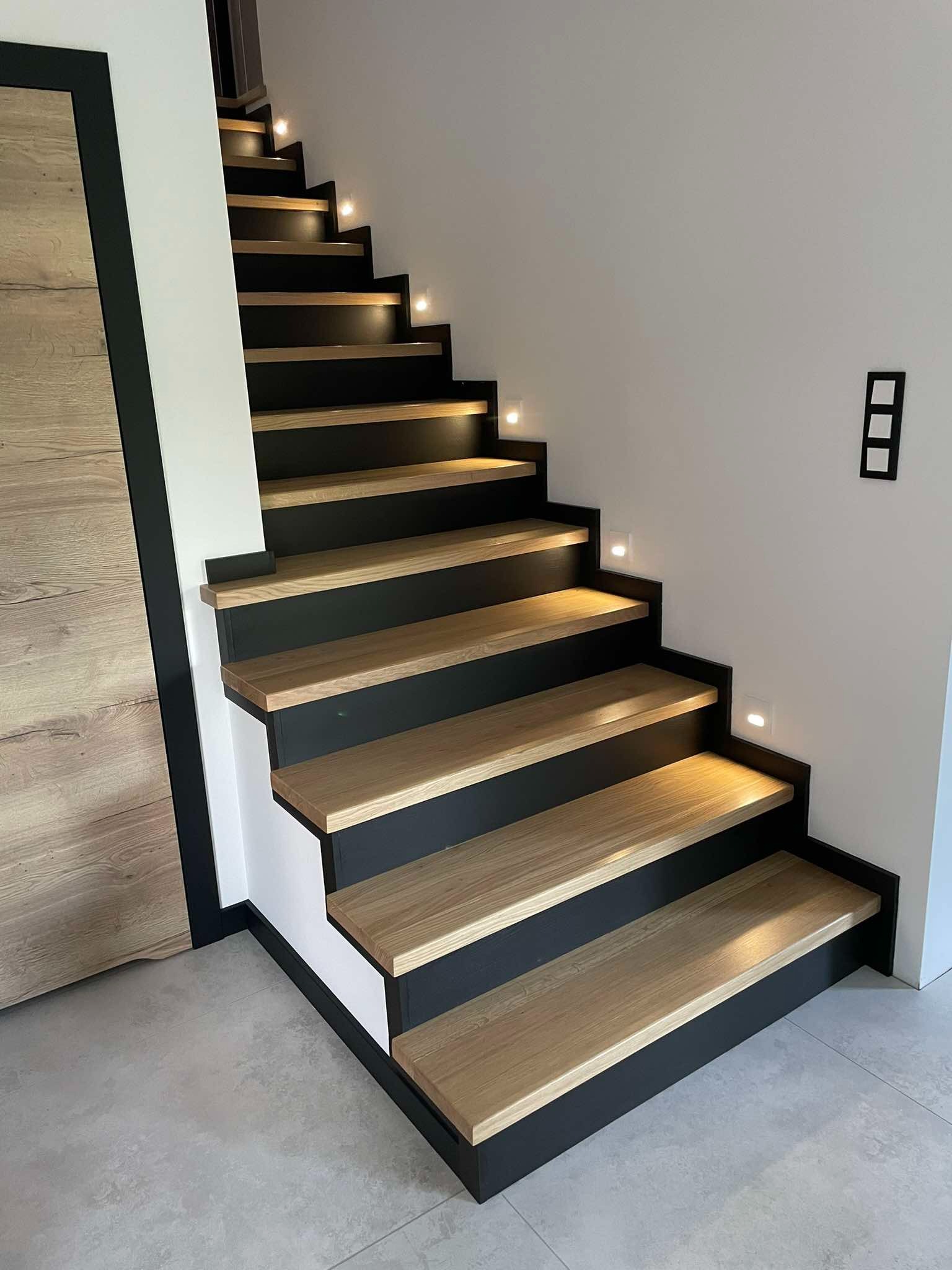Nowoczesne schody z drewnianymi stopniami i czarną konstrukcją, oświetlone punktowymi lampkami. Minimalistyczny design z drewnianymi drzwiami w tle.