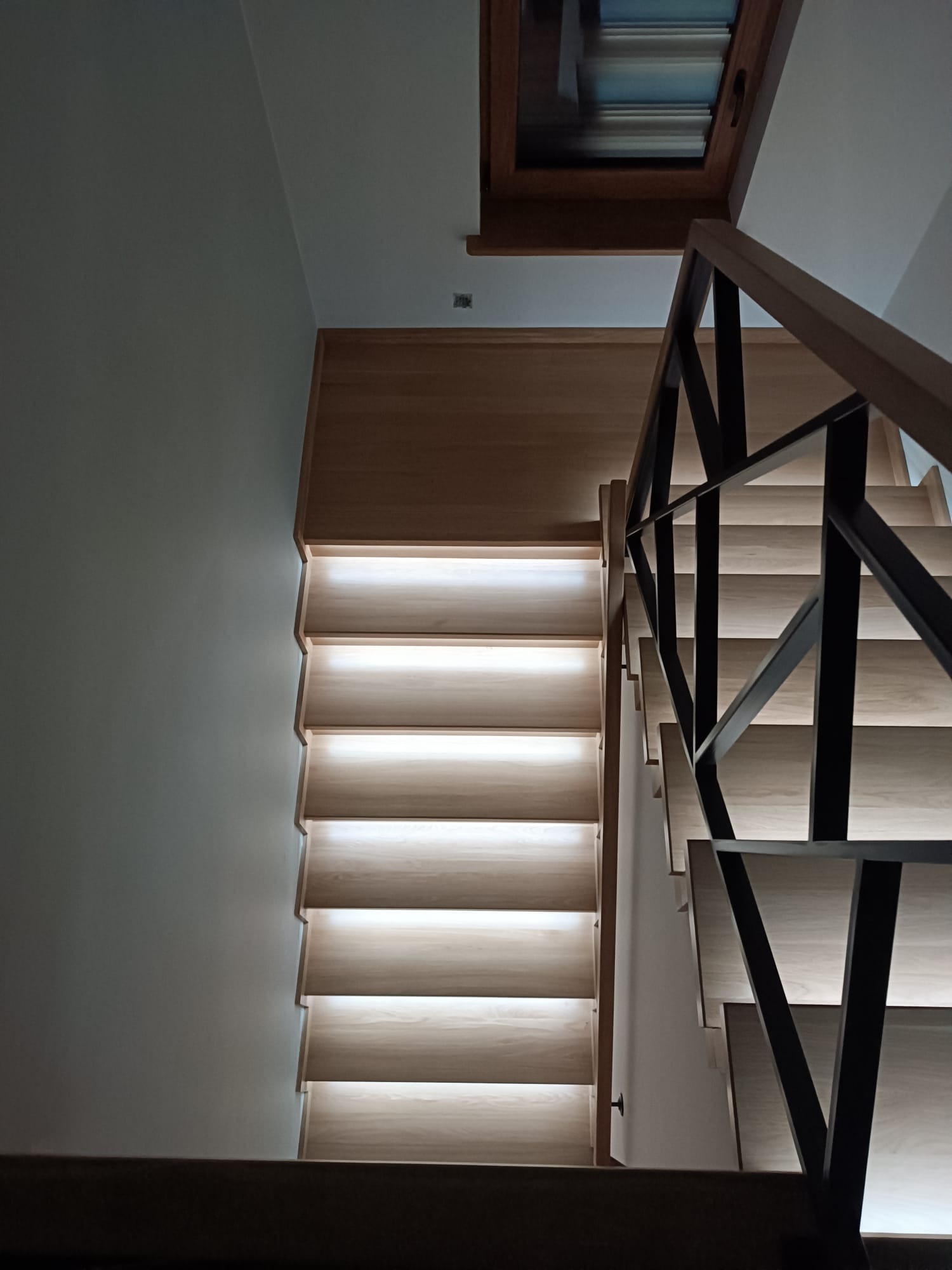 Nowoczesne, podświetlane schody z jasnego drewna, z czarną, geometryczną balustradą. Ujęcie z dołu, widoczne okno dachowe. Minimalistyczny design.