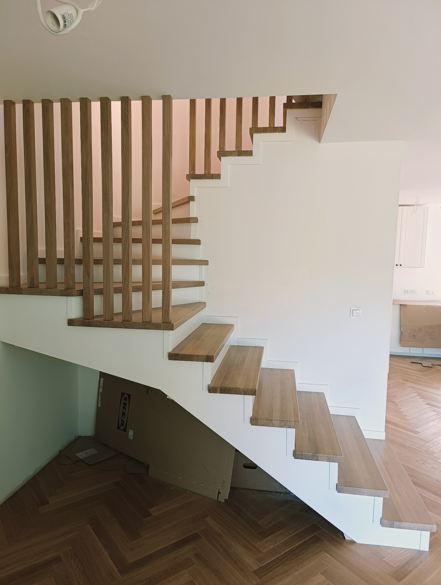 Nowoczesne schody z drewnianymi stopniami i pionową balustradą z listew, biała konstrukcja, parkiet w jodełkę na podłodze, widok z dołu.