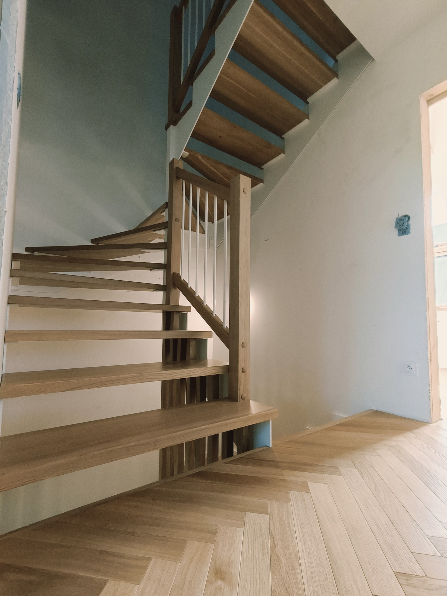 Jasne, drewniane schody spiralne z białą balustradą, widok z dołu, parkiet w jodełkę na podłodze. Minimalistyczny design, naturalne światło.