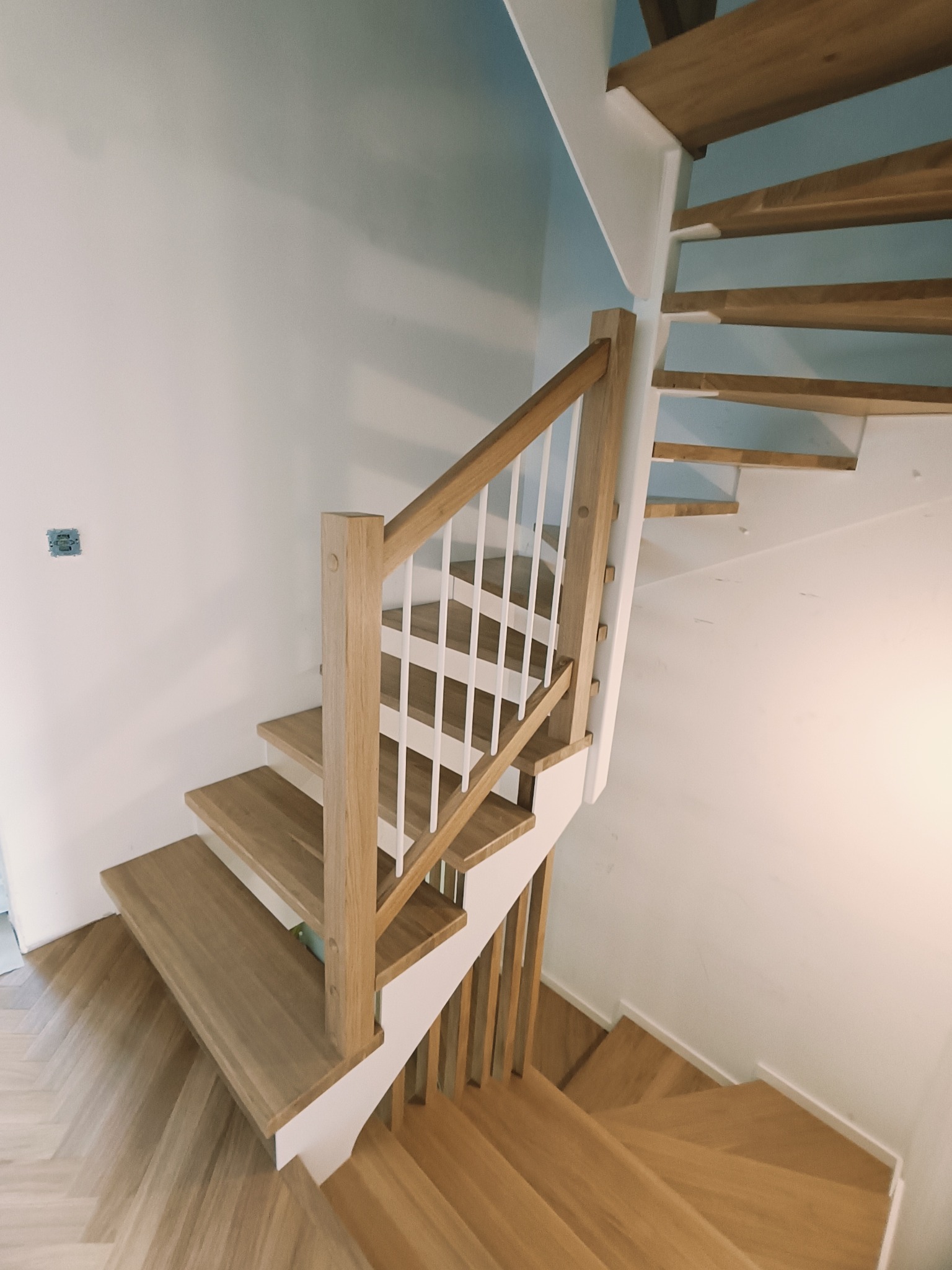 Jasne, drewniane schody spiralne z białą balustradą, widok z góry. Parkiet ułożony w jodełkę. Minimalistyczny design, nowoczesne wykończenie wnętrza.