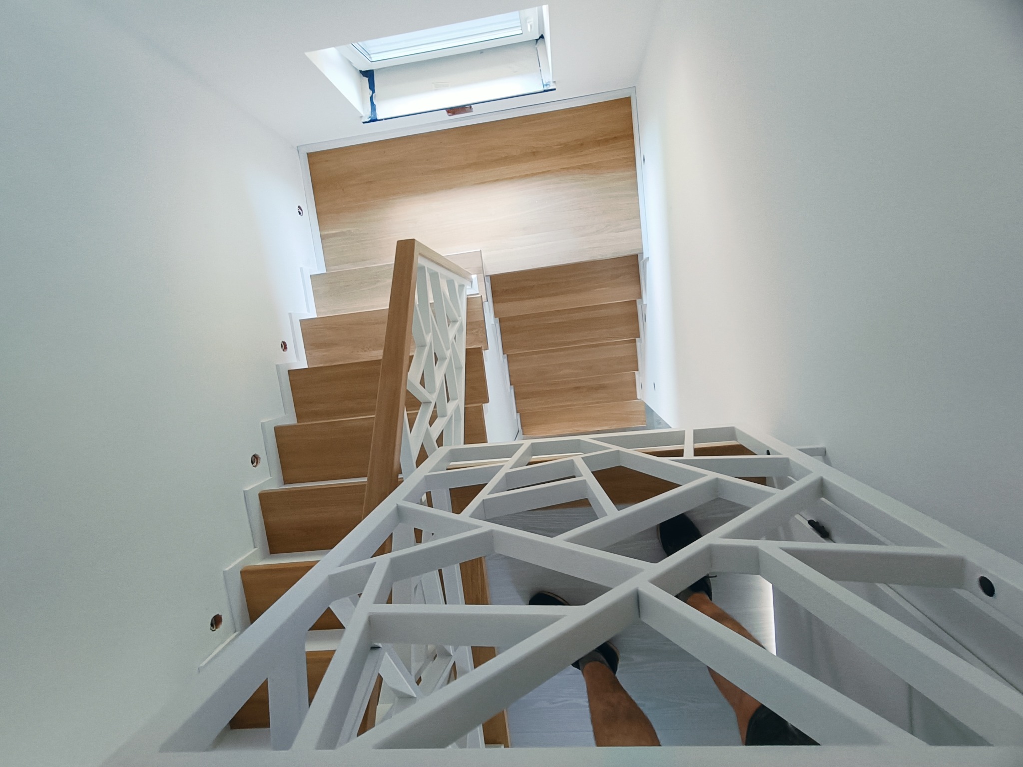 Nowoczesne schody drewniane z białą, geometryczną balustradą, widziane z góry. Naturalne światło z okna dachowego oświetla drewniane stopnie.
