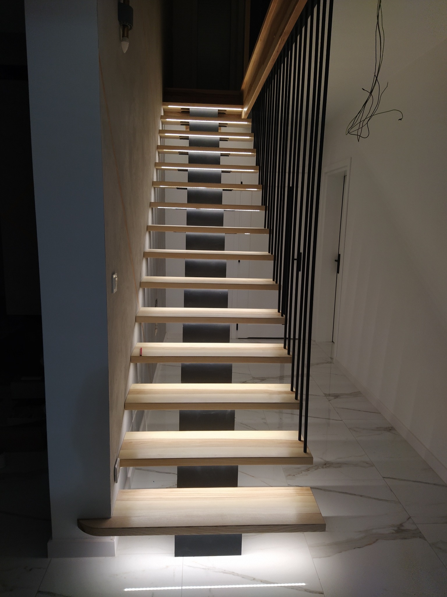 Nowoczesne schody z jasnego drewna z podświetleniem LED i czarną balustradą z pionowych prętów. Marmurowa podłoga i minimalistyczne wnętrze.