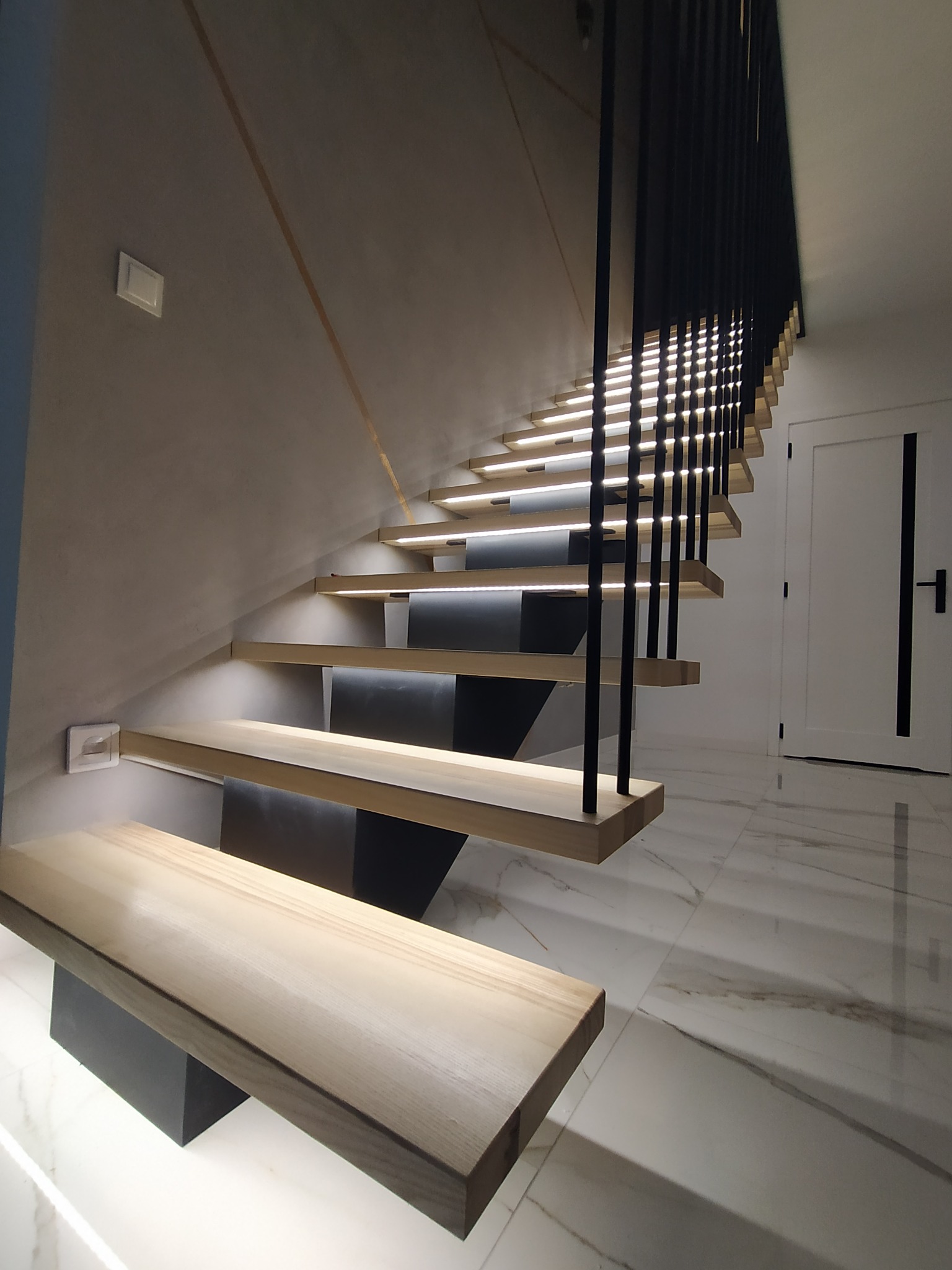 Nowoczesne schody z podświetlanymi stopniami i czarną balustradą. Minimalistyczny design, jasne drewno, marmurowa podłoga. Widok z dołu, oświetlenie LED.