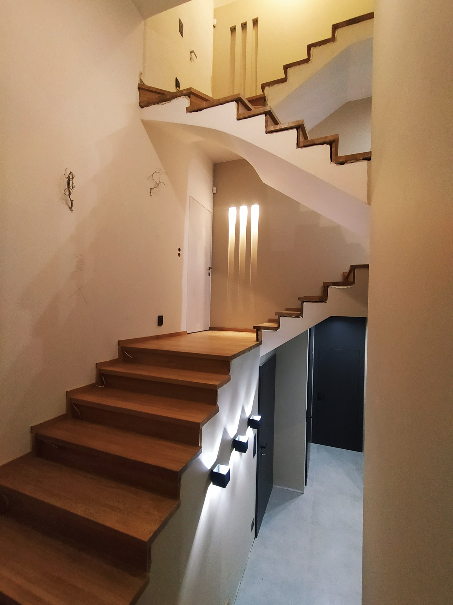 Montaż dębowych schodów w jasnym wnętrzu. Widoczne niedokończone prace wykończeniowe na ścianach i brak balustrady. Oświetlenie schodowe dodaje nowoczesnego charakteru.