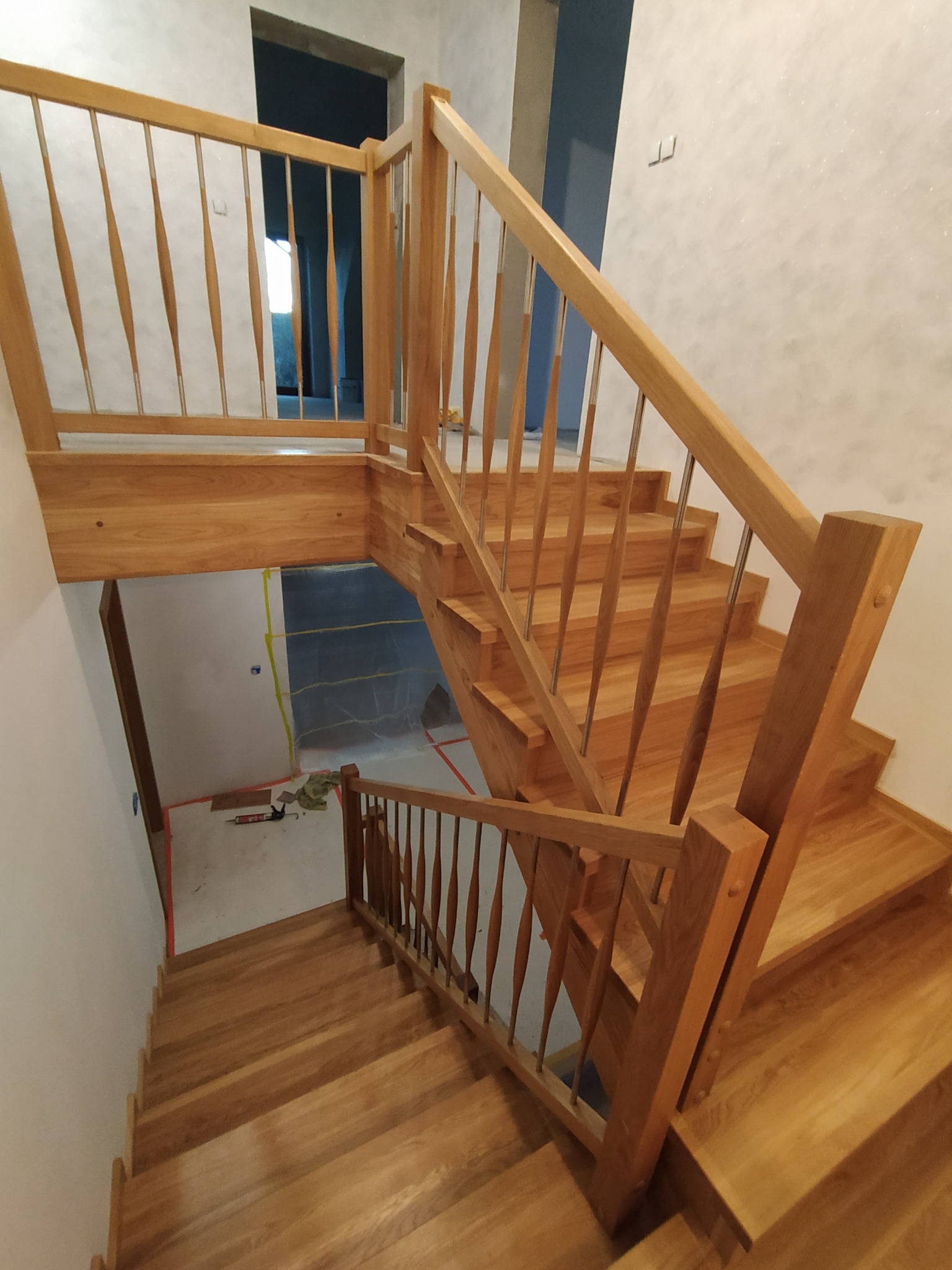 Drewniane schody z balustradą z drewnianymi szczeblami o unikalnym kształcie klepsydry, widok z góry na dół, z widocznymi narzędziami i folią ochronną na podłodze, sugerującymi etap wykończeniowy.