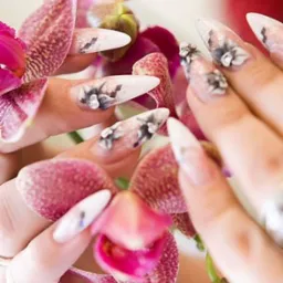 Dłonie z białymi, szpiczastymi paznokciami zdobionymi motywem kwiatowym, trzymające kwitnącą orchideę w odcieniach różu i fioletu.