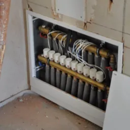 Rozdzielacz ogrzewania podłogowego w zabudowie ściennej, widoczne rury, zawory, białe termostaty i okablowanie, fragment drabiny malarskiej w lewym rogu.