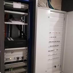 Zakład Instalatorstwa Elektrycznego Lech Różański - Rozdzielnica elektryczna z otwartymi drzwiami, widoczne zabezpieczenia i okablowanie, obok szafa rack z urządzeniami i patch panelem, część kabli prowadzona w korytach pod sufitem.
