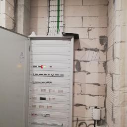 Zakład Instalatorstwa Elektrycznego Lech Różański - Otwarta szafa rozdzielcza z widocznymi bezpiecznikami i modułami sterującymi, kable w peszlach prowadzone pionowo wzdłuż surowej ściany z betonu komórkowego z widocznymi niedociągnięciami...