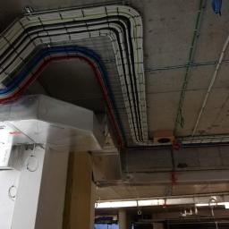 Zakład Instalatorstwa Elektrycznego Lech Różański - Widok z dołu na instalację elektryczną i wentylacyjną w surowym wnętrzu budynku, z widocznymi kablami w korytach, przewodami oraz kanałami wentylacyjnymi URSA Air.