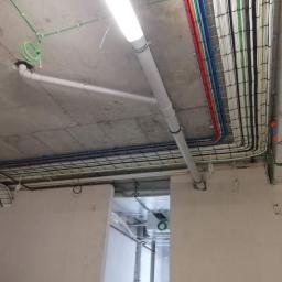Zakład Instalatorstwa Elektrycznego Lech Różański - Widok na sufit z widocznymi kablami elektrycznymi w korytkach, rurami PCV i oświetleniem liniowym w surowym, betonowym wnętrzu.