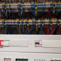 Rozdzielnica elektryczna z widocznymi bezpiecznikami, wyłącznikami i licznymi kablami oznaczonymi numerami, podłączonymi do listew zaciskowych w kolorach niebieskim, żółtym i zielonym.