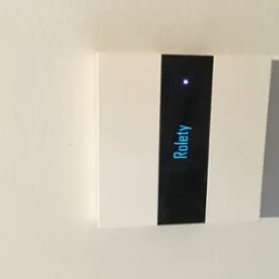 Panel sterowania roletami z podświetlanym napisem 'Rolety' zamontowany na białej, teksturowanej ścianie.